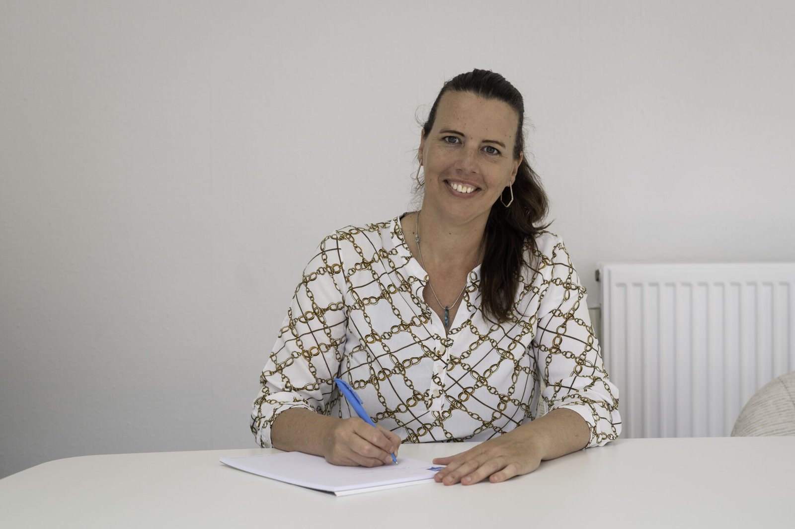 Een vrouw met lang bruin haar glimlacht naar de camera terwijl ze aan een witte tafel zit en met een blauwe pen op een notitieblok schrijft terwijl ze de Nacebelcodes doorneemt.