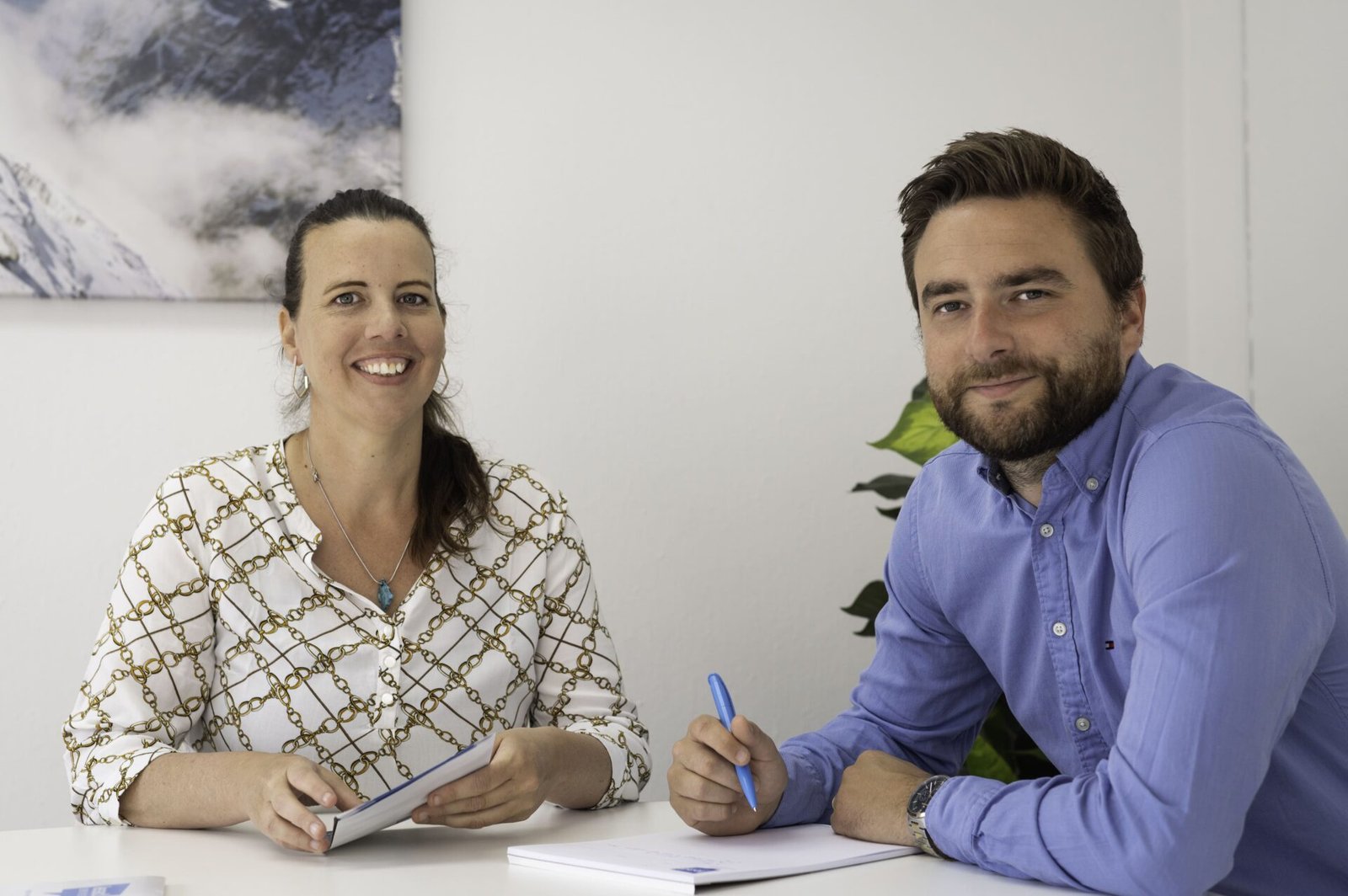 Twee mensen zitten aan een bureau met notitieboekjes en pennen en lachen naar de camera. Ze bespreken in een kantooromgeving hoe ze de Nacebel-codes voor hun bedrijf kunnen bijwerken.