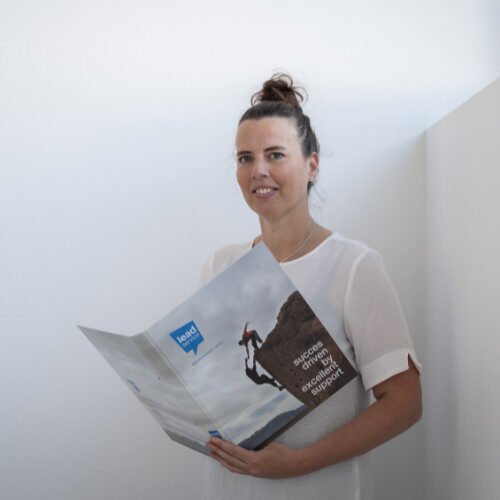 Een vrouw met bruin haar in een knot houdt een opengeslagen brochure over bergbeklimmers vast, met een tekst over succes, prestaties en hoe ze een correcte NACEBEL-registratie kan bereiken. Ze staat in een eenvoudige, lichte kamer.
